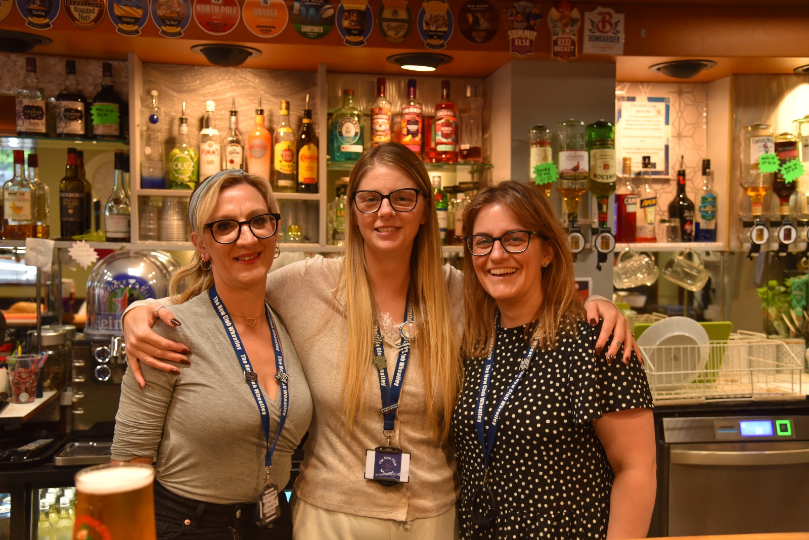 Three smiling members of bar staff in The New Club Wheatley lanyards, behind a well-stocked bar
